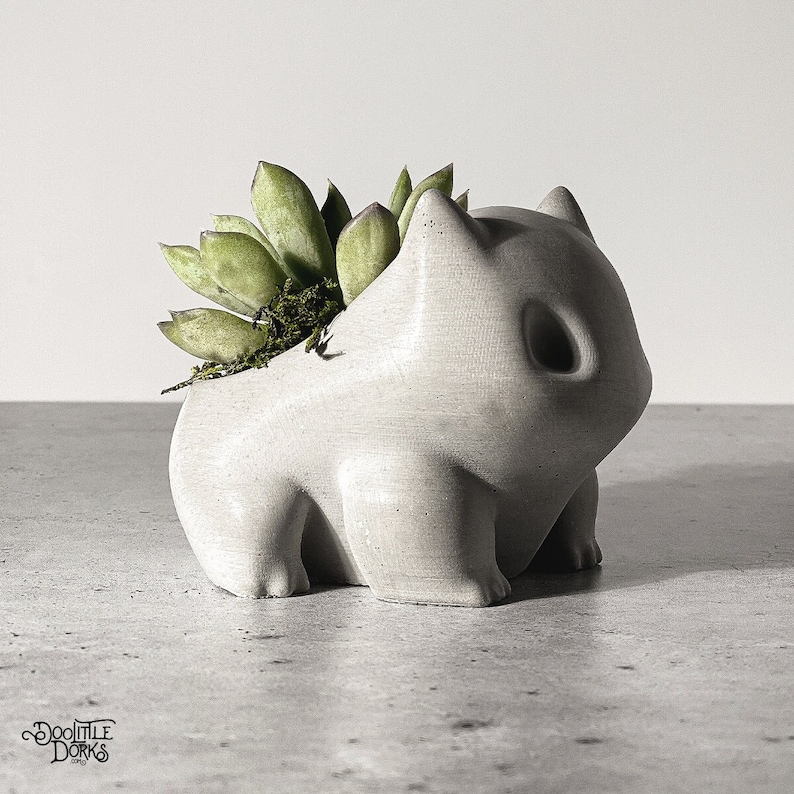 Puede incluir: Una maceta de hormig&oacute;n gris claro con forma de personaje de dibujos animados, con una cabeza grande y patas peque&ntilde;as. La maceta est&aacute; llena de suculentas verdes y musgo. La maceta est&aacute; sobre una superficie gris.