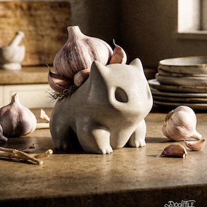 Puede incluir: Una figura de cer&aacute;mica gris claro con forma de gato, con un bulbo de ajo en la espalda. Varios dientes y bulbos de ajo est&aacute;n esparcidos alrededor de la figura sobre una encimera marr&oacute;n. La figura tiene grandes cuencas oculares.