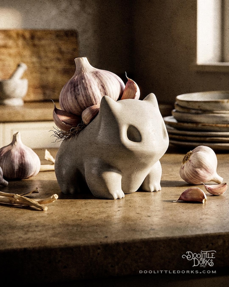 Puede incluir: Una figura de cer&aacute;mica gris claro con forma de gato, con un bulbo de ajo en la espalda. Varios dientes y bulbos de ajo est&aacute;n esparcidos alrededor de la figura sobre una encimera marr&oacute;n. La figura tiene grandes cuencas oculares.
