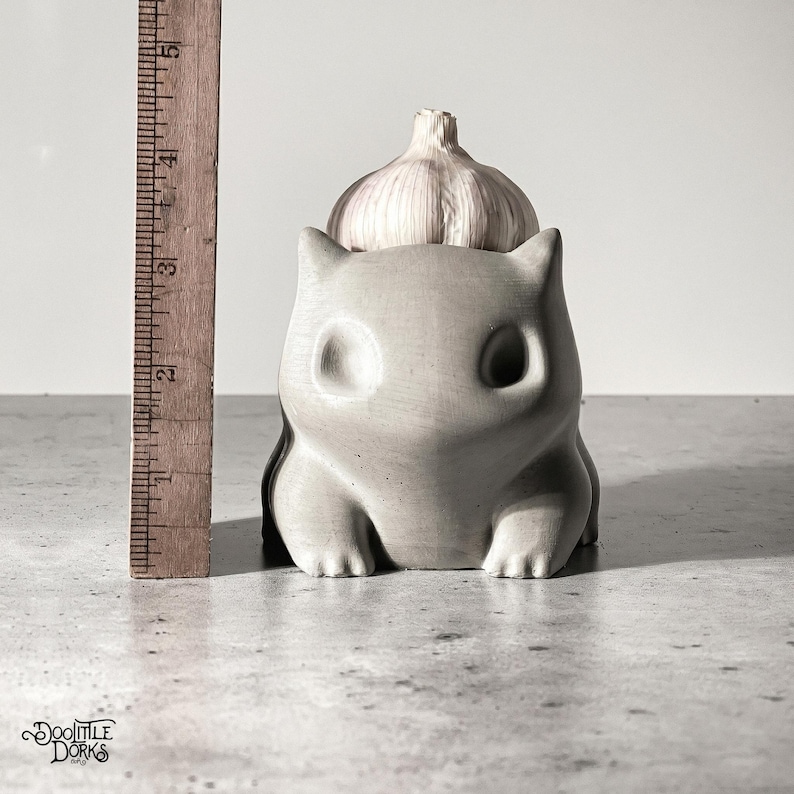May include: A grey concrete garlic keeper shaped like a cartoon animal, holding a bulb of garlic. The keeper is approximately 10 cm tall. The design has large, round eyes and a cute, squat body. The garlic sits atop the keeper.