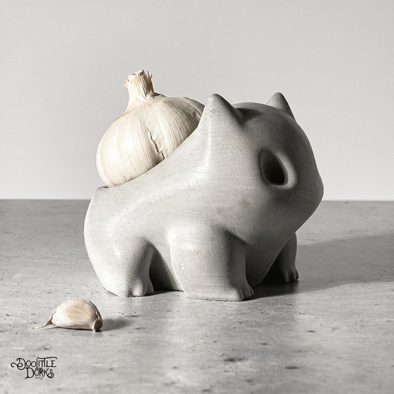 May include: A grey, concrete-effect garlic keeper shaped like a cartoon character, holding a whole garlic bulb. A single garlic clove rests on the surface. The keeper has a textured surface and a whimsical design.