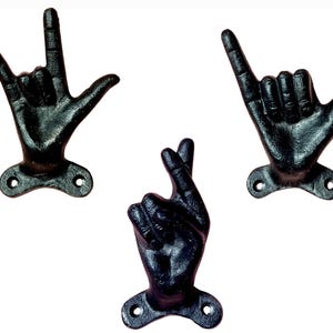 May include: Three black cast iron hand-shaped wall hooks. One hook forms the "I love you" sign, another the "hang loose" gesture, and the third has fingers crossed. Each hook has a mounting plate with two holes.