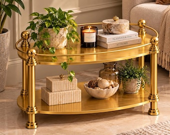 May include: A two-tier oval coffee table with a polished gold metal frame and shelves. The top shelf holds a potted plant, a candle, a lidded marble container, and two books titled The Kinfolk Home and At Home. The bottom shelf holds two stacked rectangular boxes, a bowl of decorative spheres, and a small potted plant.