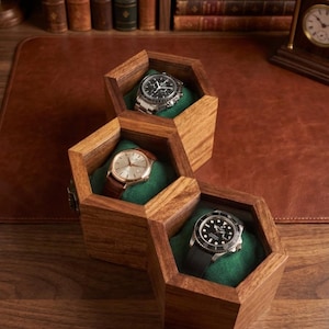 May include: A wooden watch box featuring three connected hexagonal compartments with green velvet interiors. Each compartment holds a wristwatch, including a silver chronograph, a gold-tone watch with a brown leather strap, and a black dive watch.