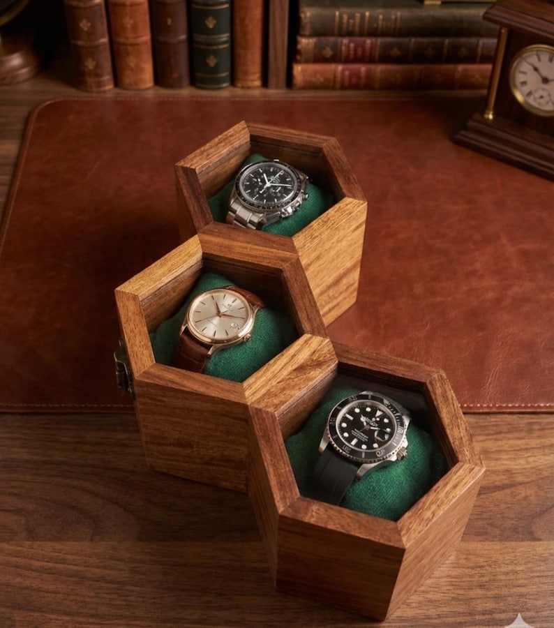 May include: A wooden watch box featuring three connected hexagonal compartments with green velvet interiors. Each compartment holds a wristwatch, including a silver chronograph, a gold-tone watch with a brown leather strap, and a black dive watch.