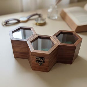 May include: A triple hexagonal wooden storage box featuring dark brown wood grain, clear glass lids, and a brass-toned metal latch on the front center compartment. The three connected containers form a honeycomb-like structure with visible green interior lining.
