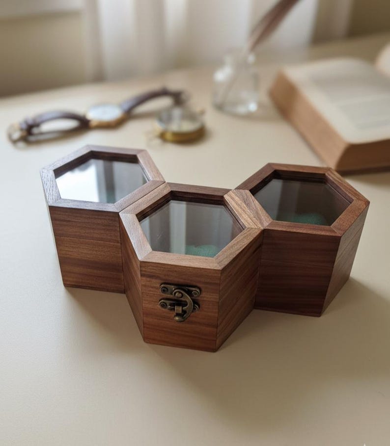 May include: A triple hexagonal wooden storage box featuring dark brown wood grain, clear glass lids, and a brass-toned metal latch on the front center compartment. The three connected containers form a honeycomb-like structure with visible green interior lining.
