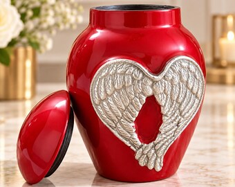 Op de afbeelding: Een glanzende rode urn met een opvallend zilverkleurig reliëf van engelvleugels in de vorm van een hart aan de voorzijde. De urn heeft een bijpassend rood deksel dat los naast de houder ligt, waardoor een zwarte binnenrand zichtbaar is.