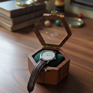 May include: A wristwatch with a silver case and a dark brown leather strap resting on a green velvet cushion inside an open hexagonal wooden box. The box features a hinged lid with a clear glass top and a small brass latch on the front edge.