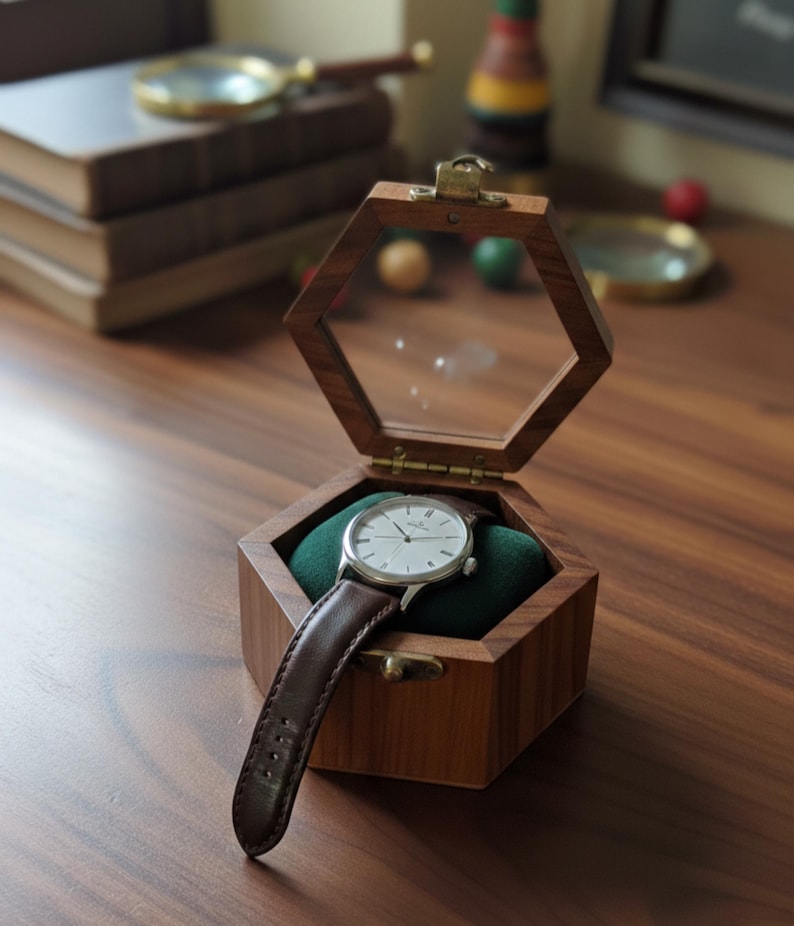 May include: A wristwatch with a silver case and a dark brown leather strap resting on a green velvet cushion inside an open hexagonal wooden box. The box features a hinged lid with a clear glass top and a small brass latch on the front edge.