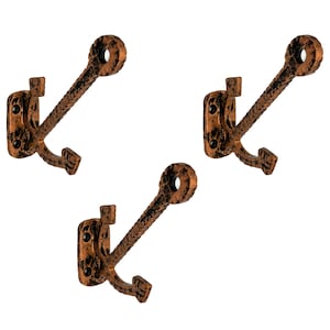 Puede incluir: Tres perchas de pared de color bronce rústico. Cada gancho presenta un diseño decorativo con un acabado texturizado. Los ganchos están montados sobre una base rectangular con dos orificios para tornillos y un solo gancho debajo.