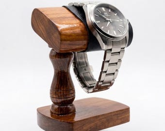May include: A silver-toned analog wristwatch with a dark grey dial and a stainless steel link bracelet, displayed on a polished dark brown wooden stand featuring a curved top cushion wrapped in black leather for secure watch placement.