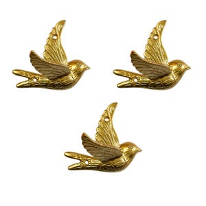 Może przedstawiać: Trzy złote metalowe dekoracje w kształcie ptaków z rozłożonymi skrzydłami. Każdy ptak ma szczegółowy wzór piór i mały otwór do zawieszenia. Ptaki są w pozycji lotu, z dziobami skierowanymi do przodu.