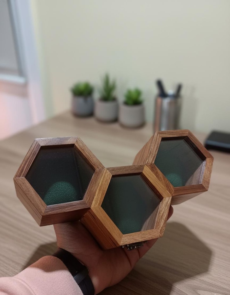 May include: A wooden display case featuring three connected hexagonal compartments with clear glass lids and dark green velvet interiors. The structure is crafted from natural brown wood with visible grain patterns, designed to hold small items securely.