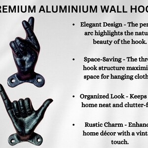 May include: Two black aluminum wall hooks shaped like hands, one with fingers crossed and the other with a shaka sign. The hooks are mounted on a white background and are designed for hanging clothes.