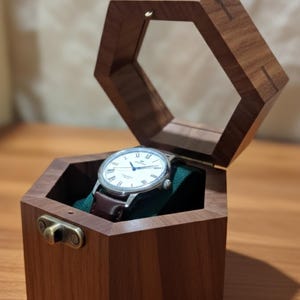 May include: A hexagonal wooden watch box with a hinged lid and brass latch, featuring a dark brown wood grain finish. Inside, a silver-cased wristwatch with a white dial and dark brown leather strap rests on a dark green velvet cushion.