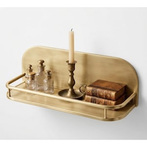 May include: A wall-mounted brass shelf with a rounded back panel and a front railing. The shelf holds three small clear glass apothecary bottles, a brass candlestick with a tall cream-colored taper candle, and two stacked antique-style books.