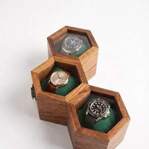 May include: Three connected hexagonal wooden watch boxes with clear glass lids. Each box features a dark green velvet interior cushion holding a different wristwatch: one with a black dial, one with a gold dial and brown strap, and one with a black bezel.