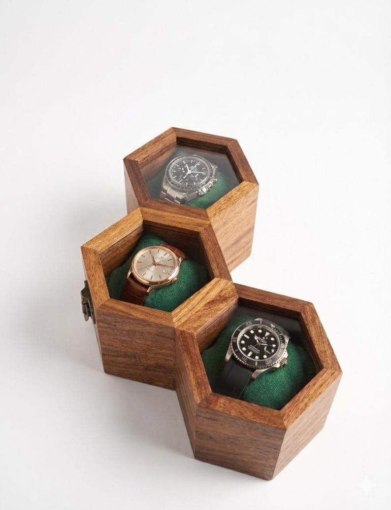 May include: Three connected hexagonal wooden watch boxes with clear glass lids. Each box features a dark green velvet interior cushion holding a different wristwatch: one with a black dial, one with a gold dial and brown strap, and one with a black bezel.