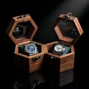 May include: Two hexagonal wooden watch boxes with hinged lids and brass-toned latches. Each box features a glass-topped lid, a dark green velvet cushion, and a silver-toned wristwatch. One watch has a blue dial, and the other has a white dial.