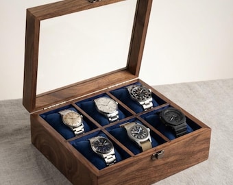 May include: A rectangular wooden watch display box with a hinged glass lid and a metal latch. The interior features six individual compartments lined with dark blue velvet, each containing a wristwatch. A small wooden tag reads Walnut & Velvet Co.