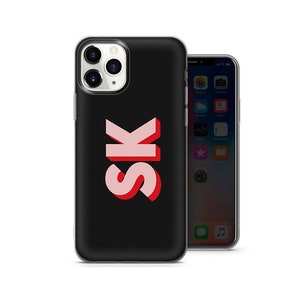 May include: Black phone case featuring the letters "SK" in pink with a red shadow. The phone case is set against a white backdrop. Another phone is visible in the background.
