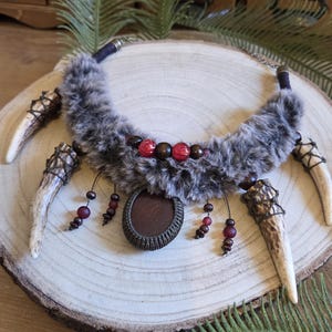 Op de afbeelding: Een unieke ketting met een harige, grijze kraag, versierd met rode en bruine kralen en een centrale bruine hanger. De ketting bevat vier geweivormige elementen en wordt gepresenteerd op een houten schijf, met een rustiek, door de natuur geïnspireerd ontwerp.
