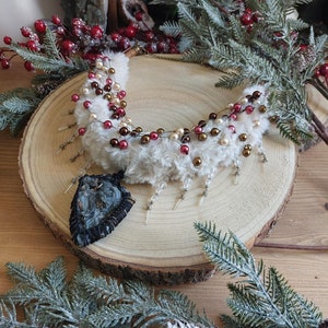 Op de afbeelding: Een witte nep bontkraag met rode, gouden en bruine kralen. De kraag wordt getoond op een houten schijf met een donkerbruine stenen hanger.