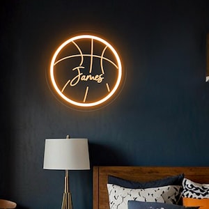 May include: A neon basketball sign with the name "James" in a script font. The sign is circular and glows with a warm, orange light. The sign is mounted on a dark blue wall above a bed with a green soccer-themed comforter.