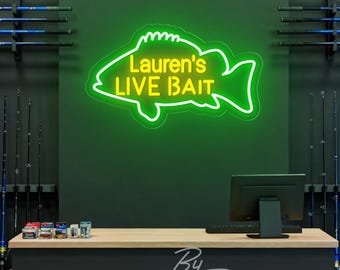 May include: A neon sign in the shape of a fish, with the text "Lauren's LIVE BAIT" in yellow against a green background. The sign is mounted on a dark wall, with fishing rods displayed on either side. A counter and computer are in the foreground.