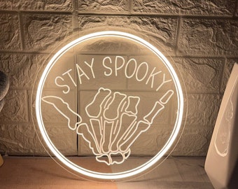 May include: A circular neon sign featuring a white glowing outline of a skeleton hand making a shaka gesture. The text STAY SPOOKY is written in a white glowing font above the hand. The entire design is enclosed within a circular neon border.