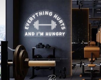 May include: A white neon sign in a gym reads "EVERYTHING HURTS AND I'M HUNGRY" with a barbell graphic. The sign is mounted on a dark gray wall. Gym equipment, including weights and benches, is visible in the background.
