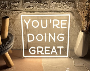 May include: A white neon sign with the words "YOU'RE DOING GREAT" in a square frame. The sign is lit up and placed against a brick wall. A white vase with dried plants is to the right.