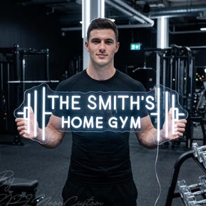 May include: A white neon sign in the shape of a barbell, with the text "THE SMITH'S HOME GYM". The sign is held by a person in a gym setting, with exercise equipment visible in the background.