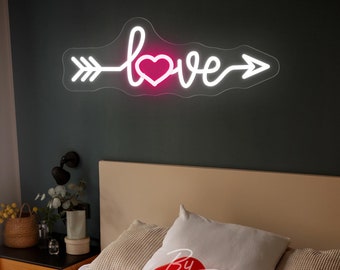 May include: White neon sign with the word "love" in a cursive font with an arrow on either side. A pink heart is in the middle of the word "love". The sign is mounted on a gray wall above a bed with white bedding and red balloons.