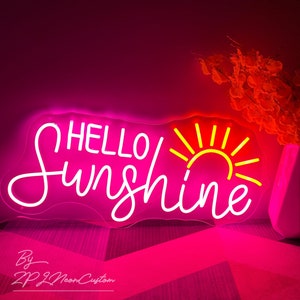 May include: A neon sign with the words "HELLO Sunshine" in white script, with a yellow sun graphic. The sign is encased in a pink outline and glows with a bright pink light. The sign is on a pink surface.