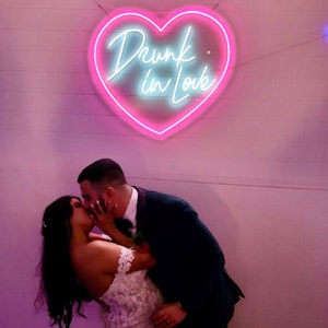 May include: A neon sign in the shape of a heart with the words "Drunk in Love" in cursive script. The sign is pink with a light blue inner glow. A couple is kissing in front of the sign.