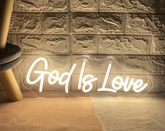 May include: A white neon sign displaying the words "God Is Love" in a cursive font. The sign is illuminated against a textured brick wall background. The words "By ZP Neon Custom" are written below the sign.