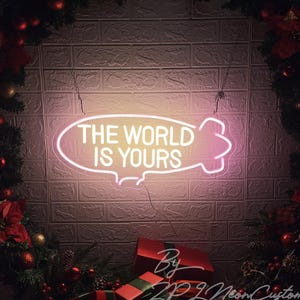 Puede incluir: Un letrero de neón con la frase "THE WORLD IS YOURS" en blanco, dentro de una forma de bocadillo. El letrero emite un suave brillo rosa, sobre una pared de ladrillos. Decoraciones navideñas festivas enmarcan el letrero.
