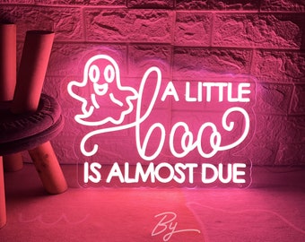 May include: A neon sign featuring a white ghost graphic and the text A LITTLE boo IS ALMOST DUE. The sign emits a bright white light against a pink-lit brick wall background, creating a vibrant display with clear, legible lettering and a cartoon ghost.