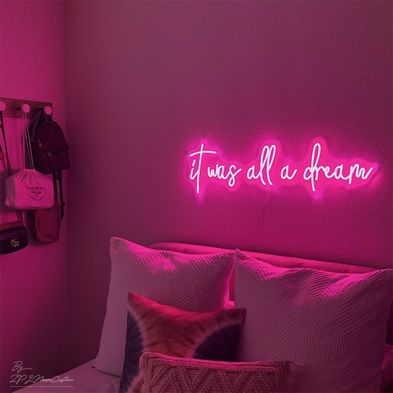 May include: A neon sign in bright pink script reads "it was all a dream" on a pink wall. The sign is above a bed with white pillows and a patterned throw pillow. A wall-mounted rack holds several bags.