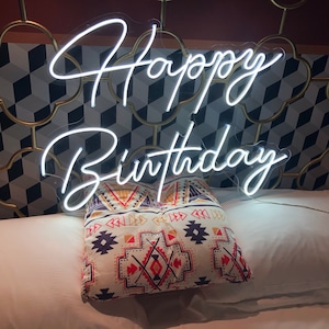 May include: A white neon "Happy Birthday" sign is displayed above a decorative pillow with a geometric pattern. The pillow is resting on a white bed. The background features a patterned wall.