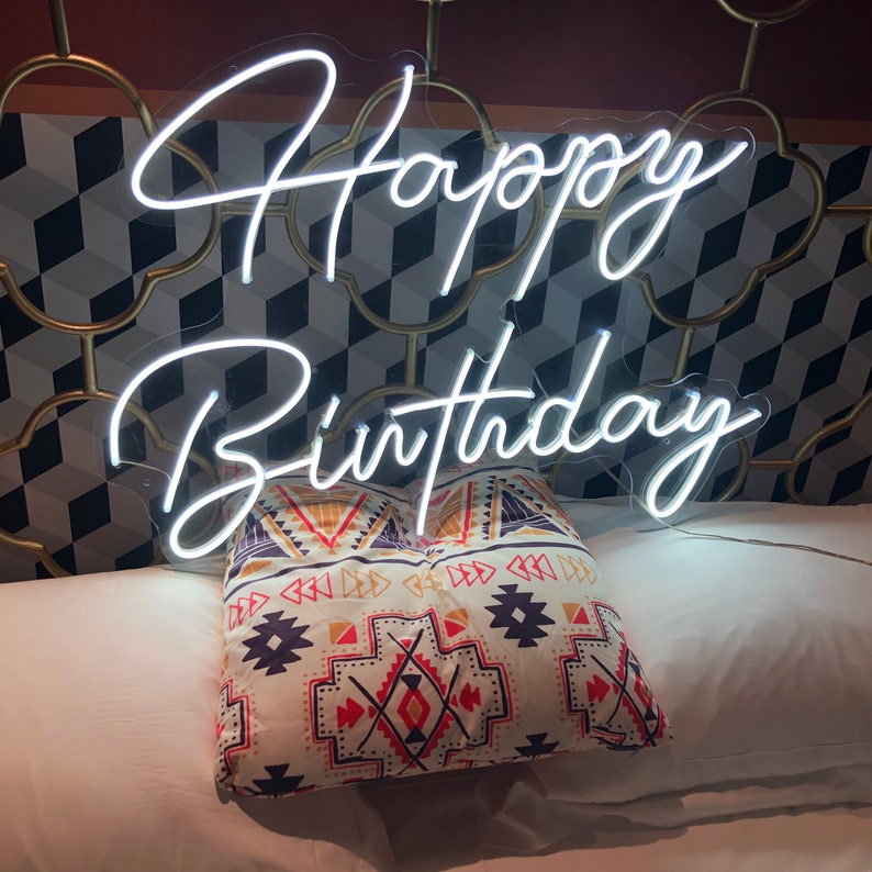 May include: A white neon "Happy Birthday" sign is displayed above a decorative pillow with a geometric pattern. The pillow is resting on a white bed. The background features a patterned wall.