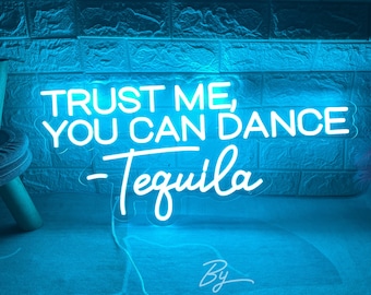 May include: A neon sign in bright blue with the text "TRUST ME, YOU CAN DANCE - Tequila". The sign is mounted on a textured blue brick wall. The sign also includes the text "By ZP Neon Custom".