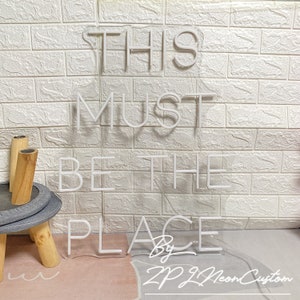 Op de afbeelding: Wit neonbord met de tekst "THIS MUST BE THE PLACE" tegen een witte bakstenen muur. Het bord bevat de tekst "By ZP ZL Neon Custom". Het bord is verlicht.