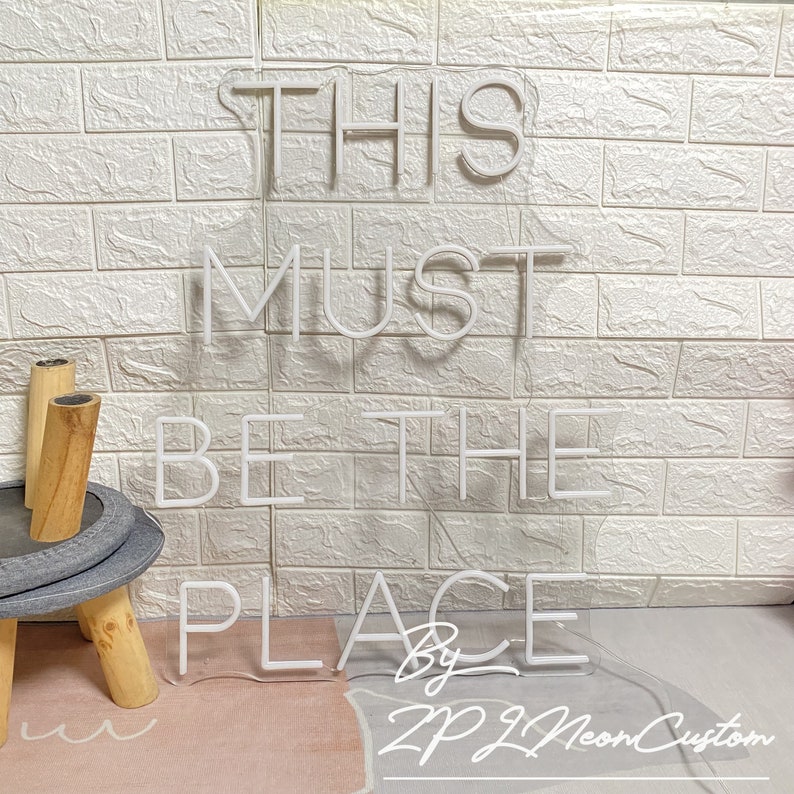 Op de afbeelding: Wit neonbord met de tekst "THIS MUST BE THE PLACE" tegen een witte bakstenen muur. Het bord bevat de tekst "By ZP ZL Neon Custom". Het bord is verlicht.