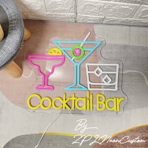 May include: A neon sign with a clear backing features cocktail glasses in pink, blue, and white, with the words "Cocktail Bar" in yellow. The sign also includes the text "By ZP Neon Custom".