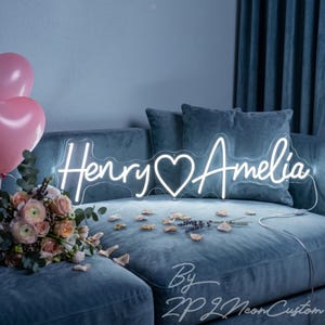 May include: A custom neon sign featuring the names Henry and Amelia in a white cursive script with a heart symbol in the center. The sign is illuminated and displays the text Henry and Amelia. It is positioned on a blue sofa with flowers.