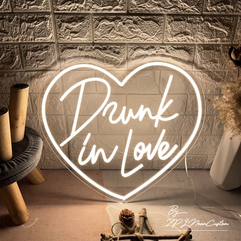 May include: A white neon sign in the shape of a heart with the words "Drunk in Love" in cursive script. The sign is illuminated against a textured brick wall background. The sign is a decorative item.