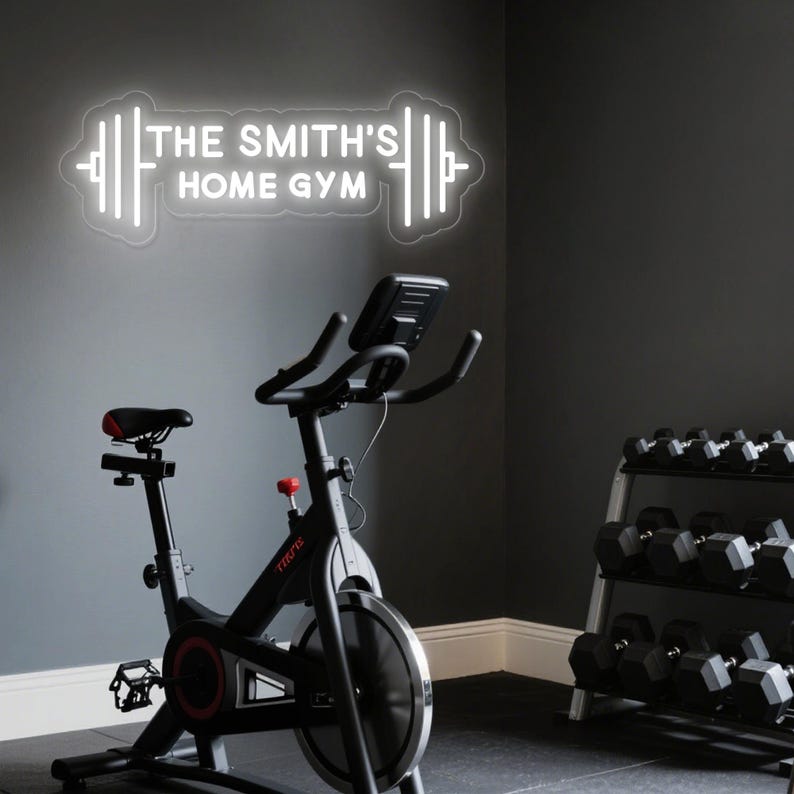 May include: A home gym setup featuring a black exercise bike with a red seat and a rack of dumbbells. A white neon sign on the wall reads "THE SMITH'S HOME GYM" in a barbell shape, creating a modern fitness space.